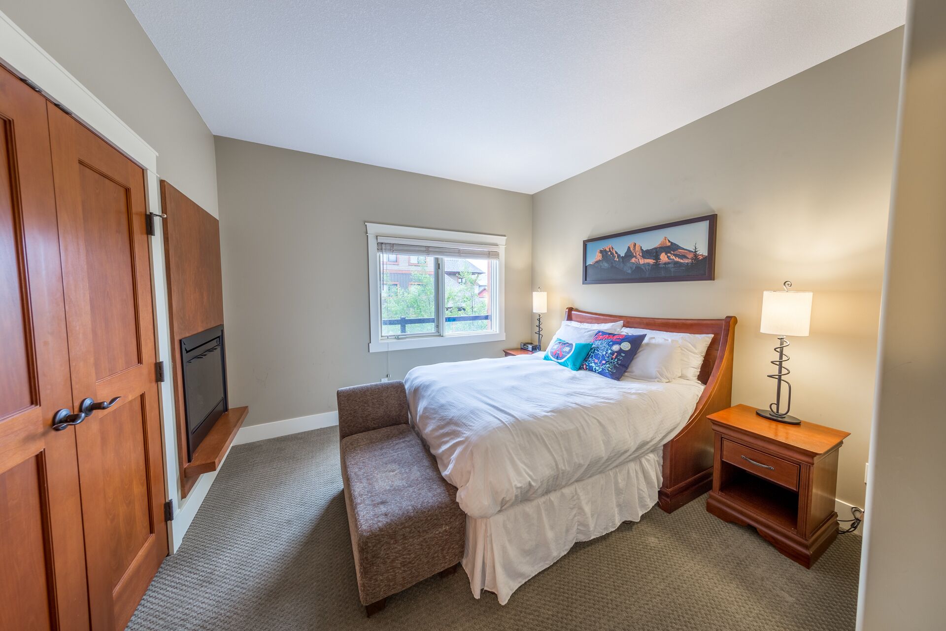 A bedroom with a bed, bench, nightstand, lamp, window, and a painting on the wall.