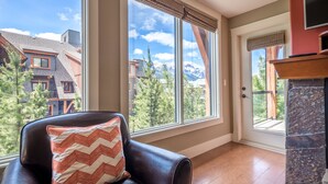 A room with a large window offering a mountain view, a fireplace, and a leather chair with a patterned pillow. - Mountain Views - Canmore Luxury Condo in Solara Resort - Steps to restaurants (Canmore)