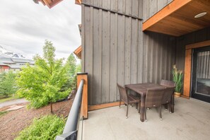 Un balcón con mesa y sillas, baranda de madera, y vistas a un jardín y edificios al fondo.