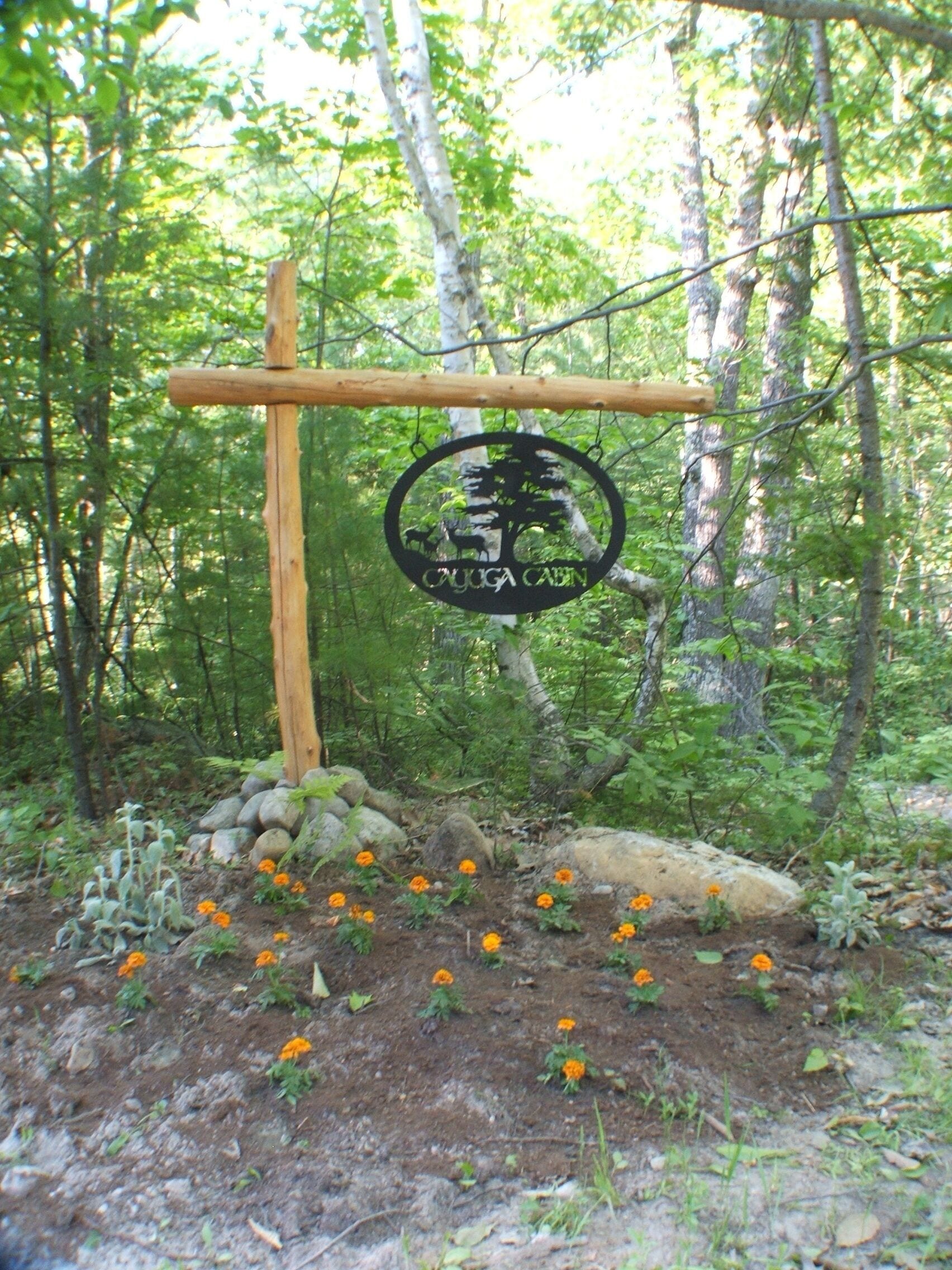 A wooden sign with a circular emblem in a forested area with rocks and orange flowers.
