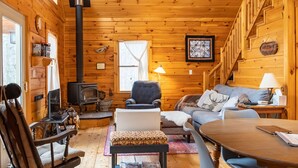 A cozy log cabin interior with a fireplace, wooden walls, a sofa, a dining table, and a staircase. - Secluded, Furnished, Log Home, Close To Many Adirondack Attractions. (Jay)