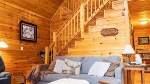 A wooden cabin interior with a staircase, a sofa with a deer pillow, a welcome sign, and a lamp. - Secluded, Furnished, Log Home, Close To Many Adirondack Attractions. (Jay)