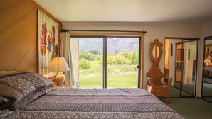 A bedroom with a large bed, a nightstand, a lamp, a wooden chair, a mirror, and a sliding glass door leading to a balcony with a mountain view. - Deluxe CREEKSIDE Condo w/SPECTAULAR Mountain Views (Mammoth Lakes)