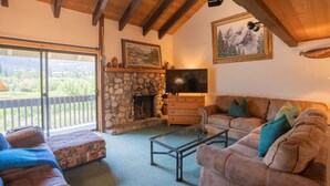 A cozy living room with a stone fireplace, a flat-screen TV, and a large window overlooking a scenic landscape. - Deluxe CREEKSIDE Condo w/SPECTAULAR Mountain Views (Mammoth Lakes)