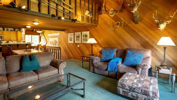 A cozy living room with leather sofas, a glass coffee table, and a patterned ottoman. A staircase with wooden railings and a chandelier is visible in the background. - Deluxe CREEKSIDE Condo w/SPECTAULAR Mountain Views (Mammoth Lakes)