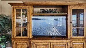 Un meuble de rangement en bois, conçu pour accueillir un téléviseur, sur lequel est diffusée une image de bateau sur l’eau. Il est entouré d’armoires et d’étagères à portes vitrées.