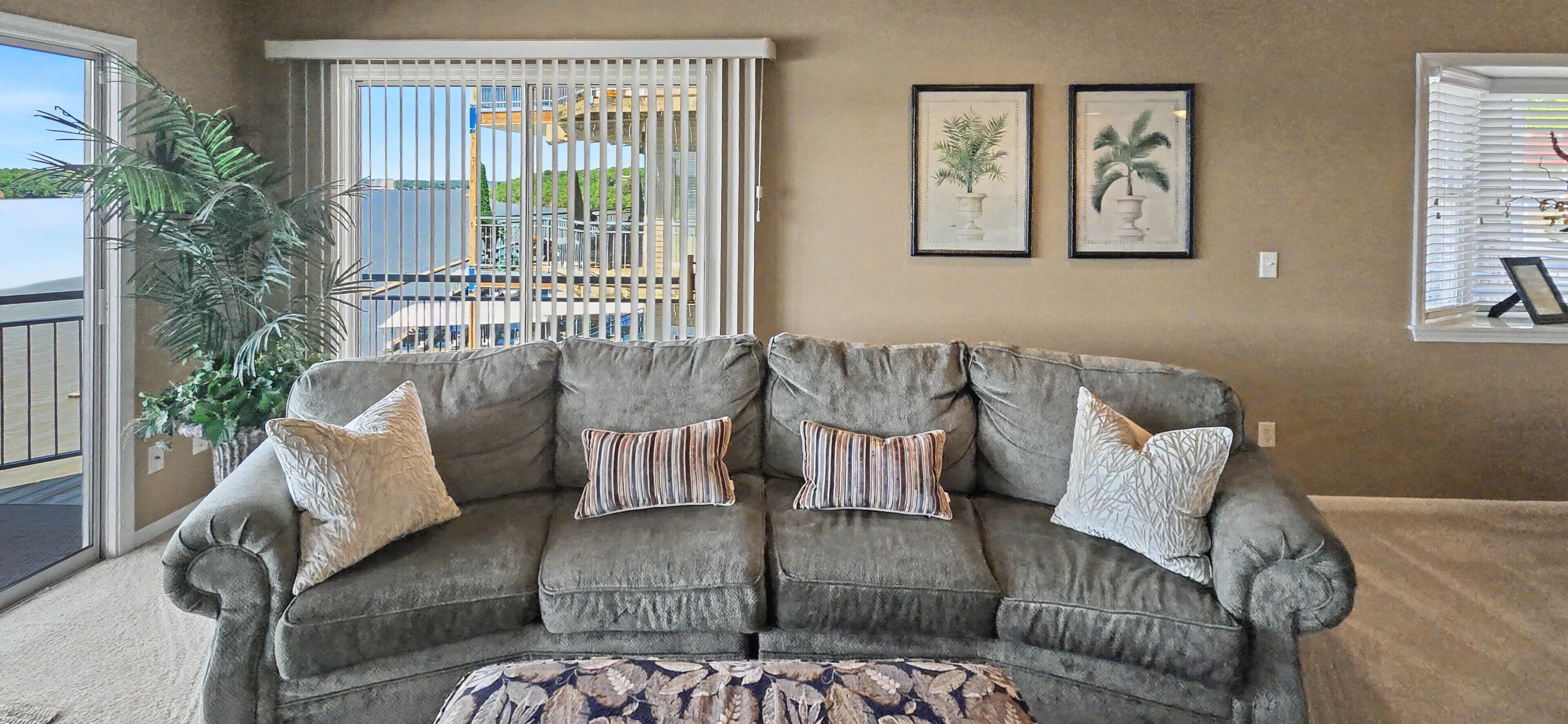 A gray sectional sofa with striped and textured pillows, a patterned ottoman, and a view of a balcony with a railing and a palm tree.