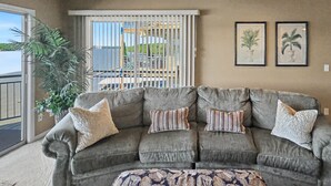 Un canapé d’angle gris avec des coussins à rayures et à motifs, un pouf à motifs et une vue sur un balcon doté d’une balustrade et d’un palmier.