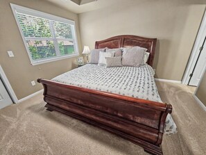 Quarto com cama de madeira, paredes bege, janela com persianas, mesa de cabeceira com abajur e piso coberto por carpete.
