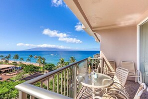 A balcony with a glass table and chairs, overlooking a beach with palm trees and the ocean.