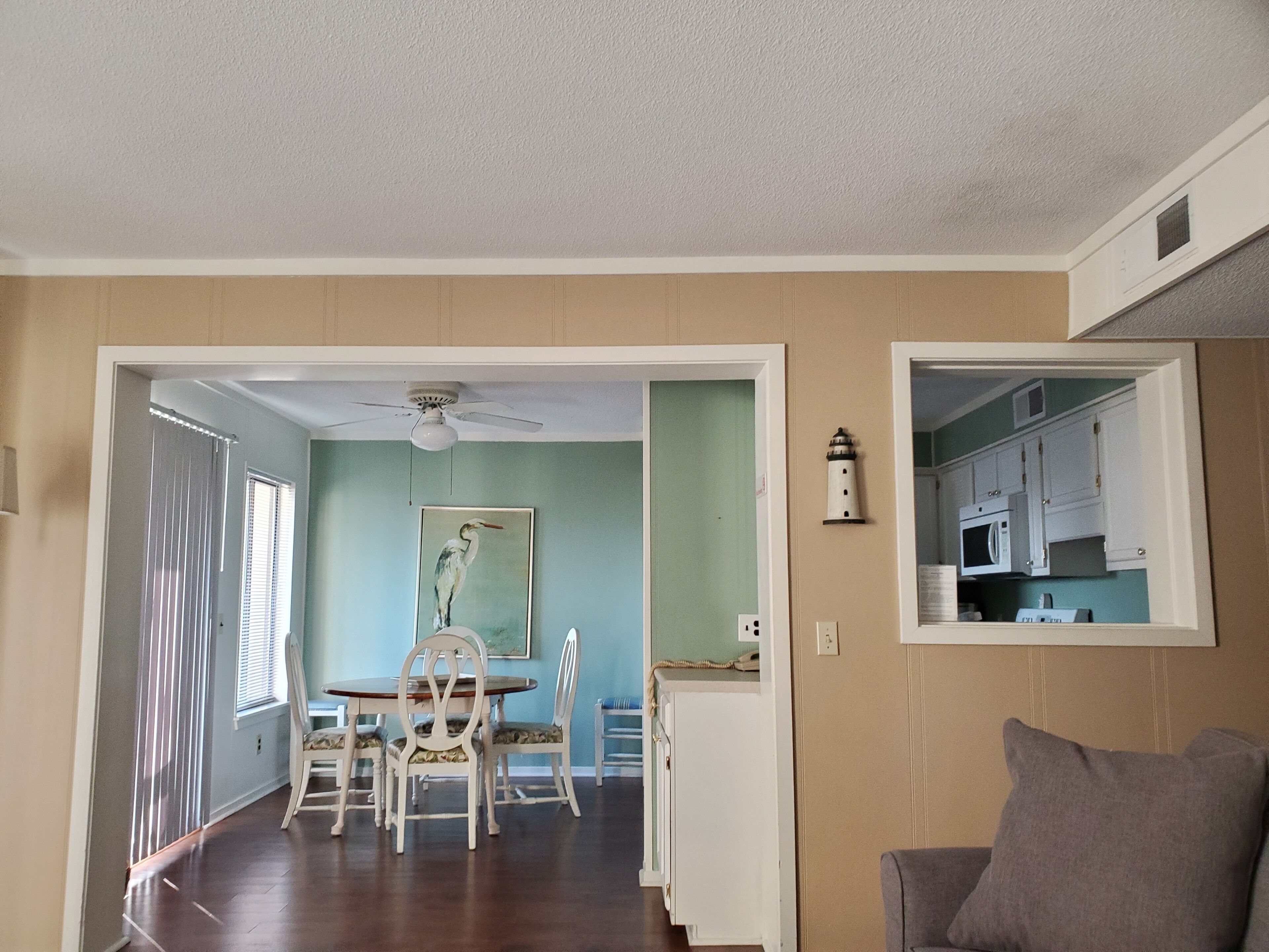 A dining area with a table and chairs, a ceiling fan, a painting, and a kitchen visible through an open door.