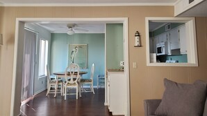 A dining area with a table and chairs, a ceiling fan, a painting, and a kitchen visible through an open door.