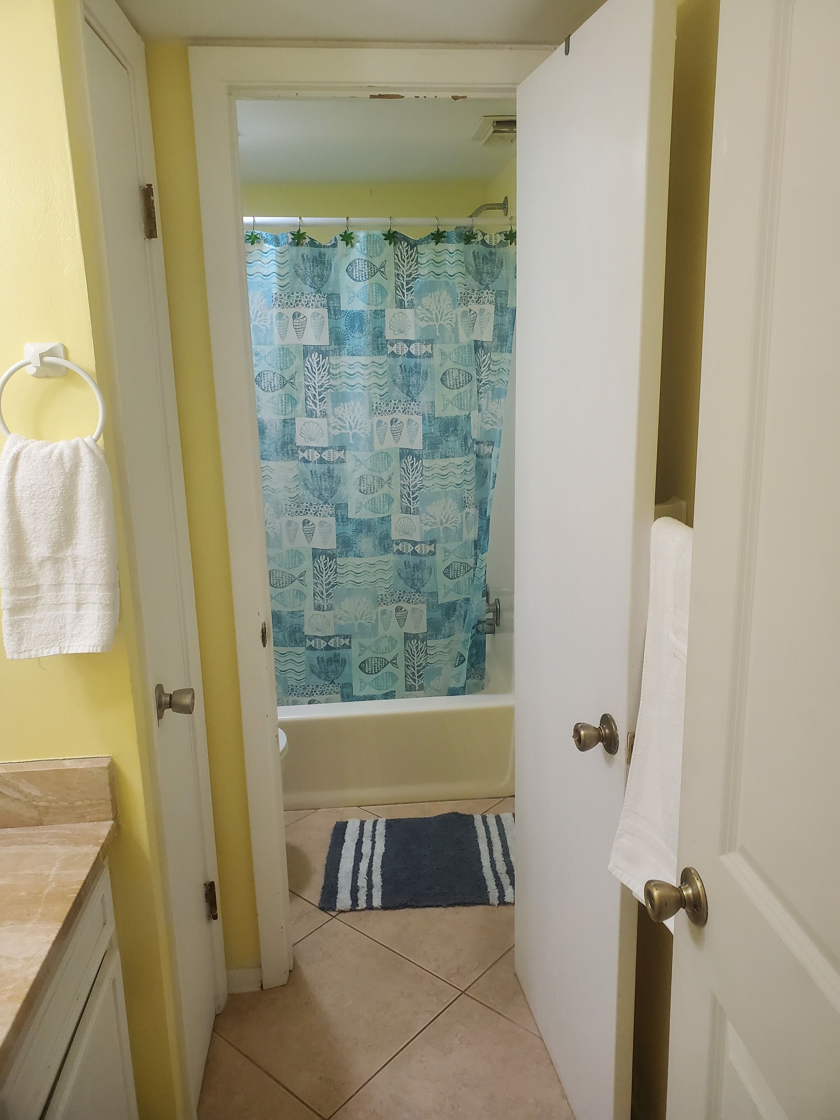 A bathroom with a shower curtain featuring a fish pattern, a white towel hanging on a ring, and a striped bath mat on the floor.