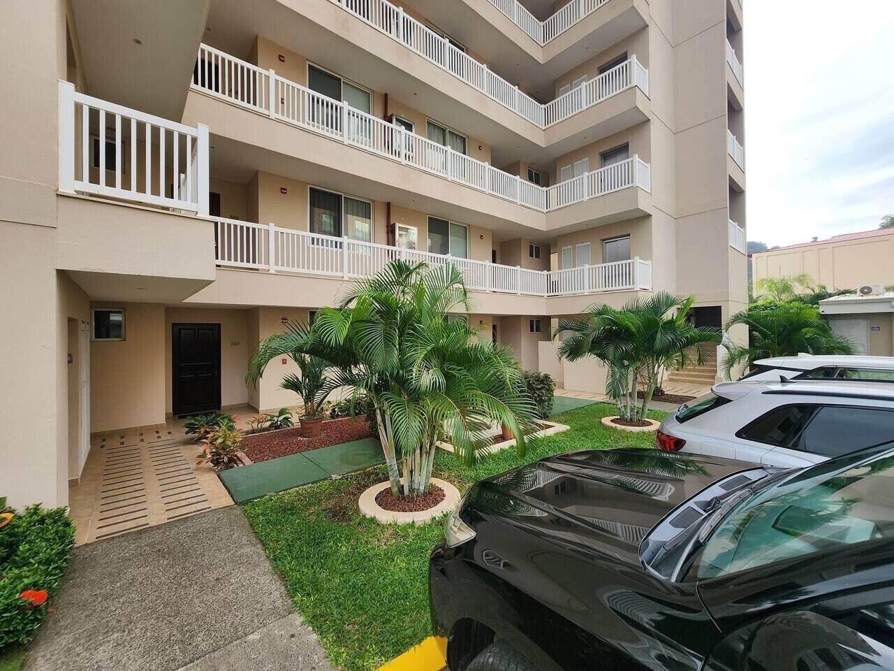 Um edifício de vários andares com varandas, área de estacionamento e um pequeno jardim com palmeiras.