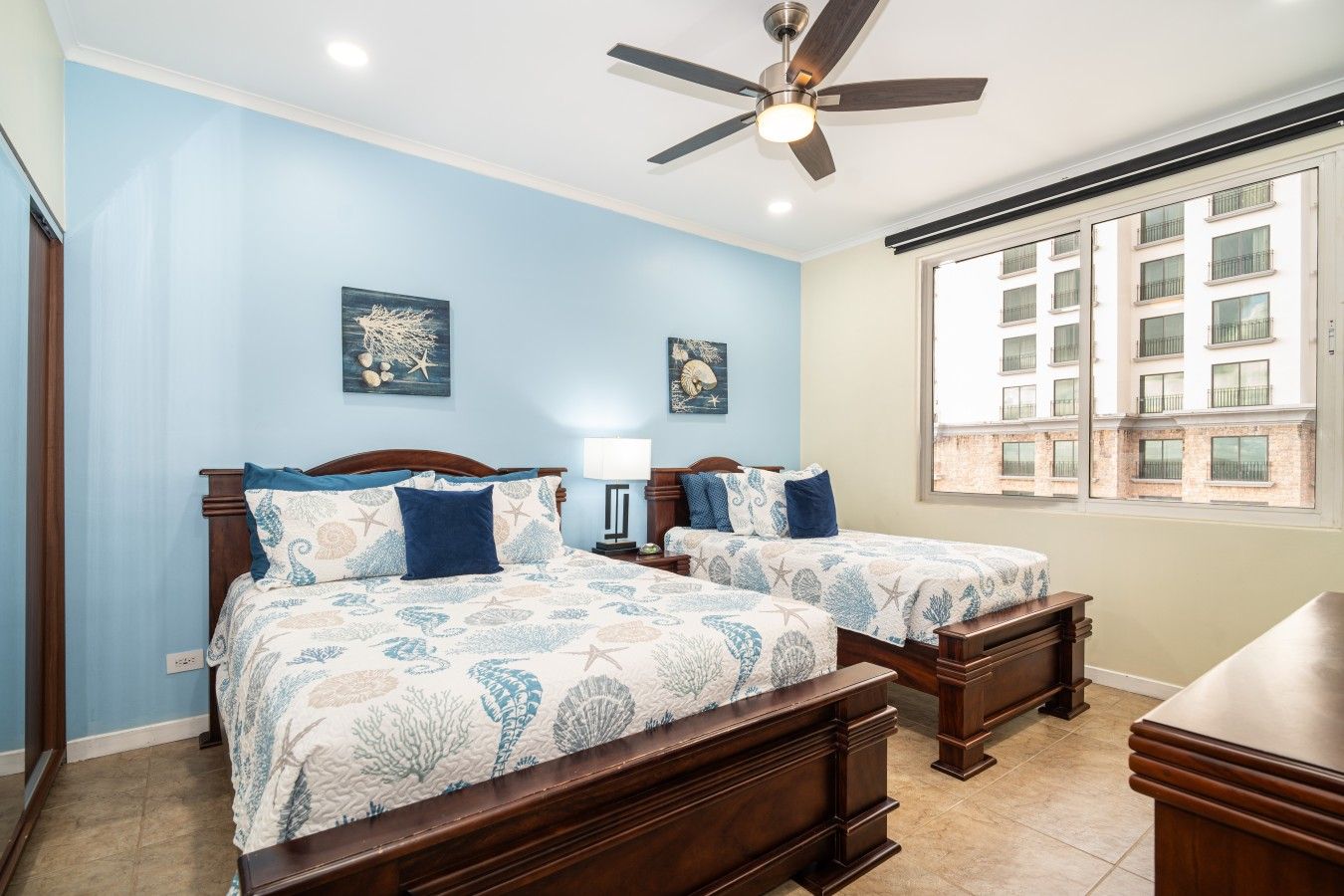 Quarto com duas camas, ventilador de teto e janela com vista para edifícios.