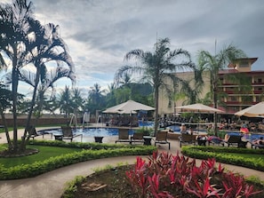 Zona de piscina en un resort con sillones de playa y sombrillas, rodeada de palmeras y un edificio al fondo.