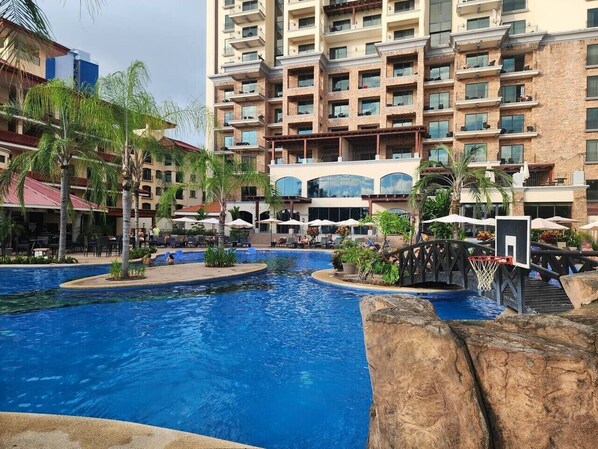 A swimming pool area with a basketball hoop, palm trees, and a multi-story building in the background.