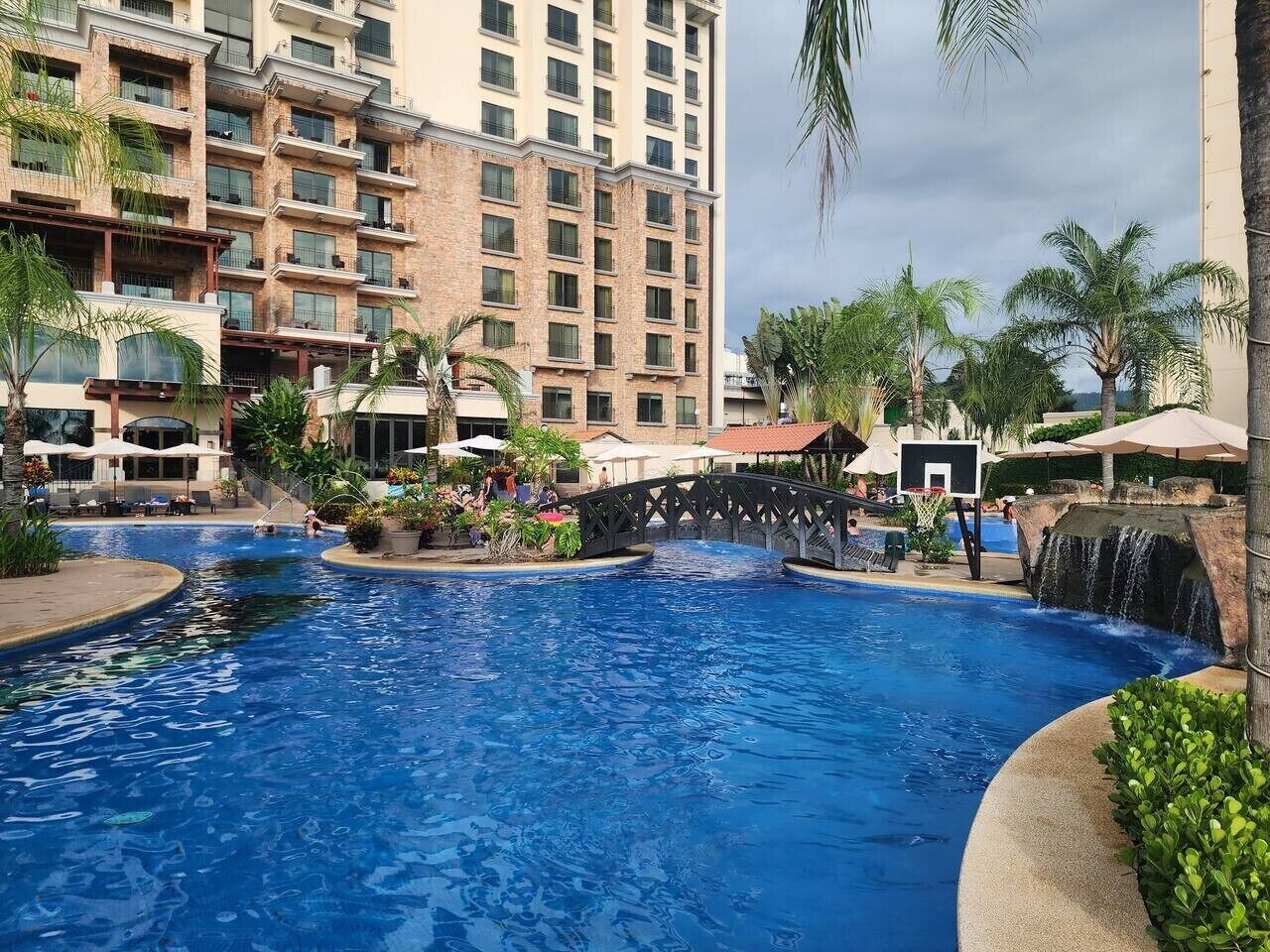 Una piscina con un puente, palmeras y un edificio de varios pisos al fondo.