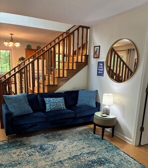 A living room with a blue sofa, a round mirror, a small table with a lamp, and a wooden staircase.