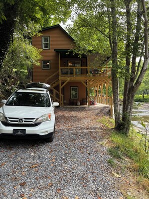 A two-story wooden cabin with a gravel driveway and a white car parked in front.