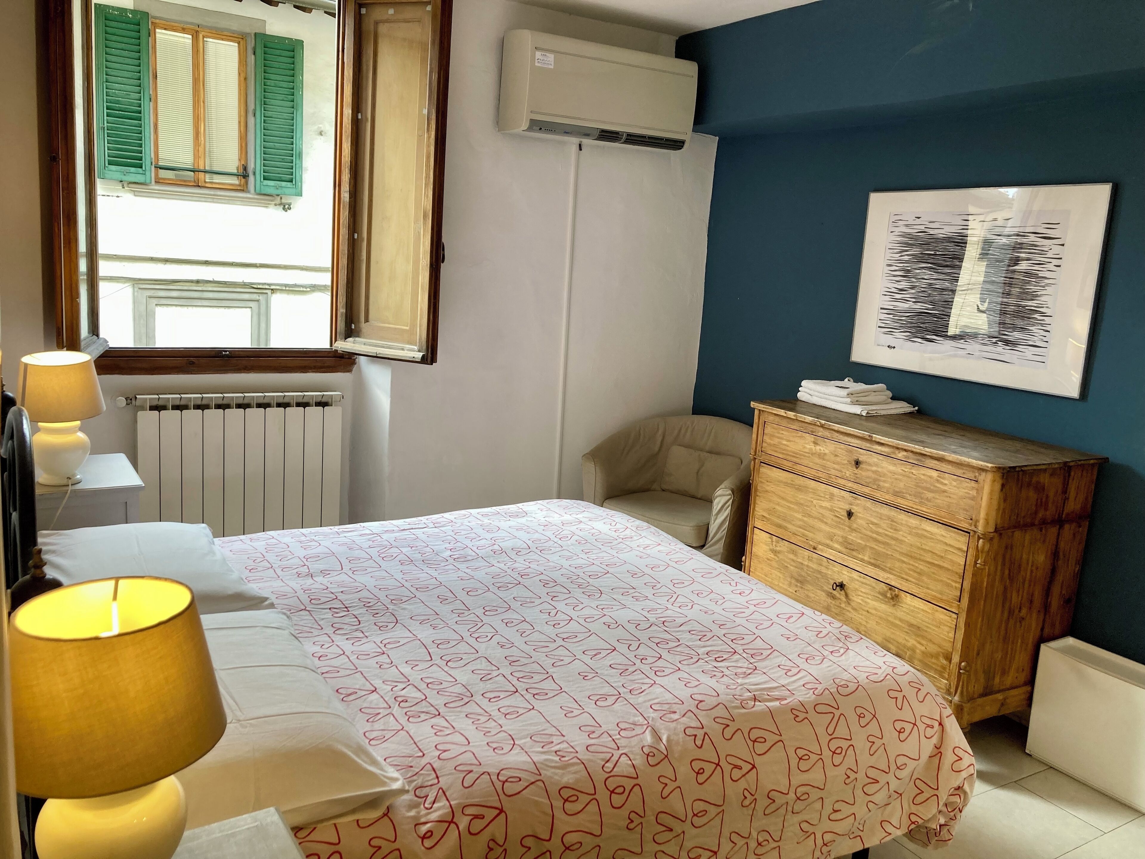 A bedroom with a bed, a wooden dresser, a radiator, an air conditioner, a window with shutters, and a wall-mounted artwork.