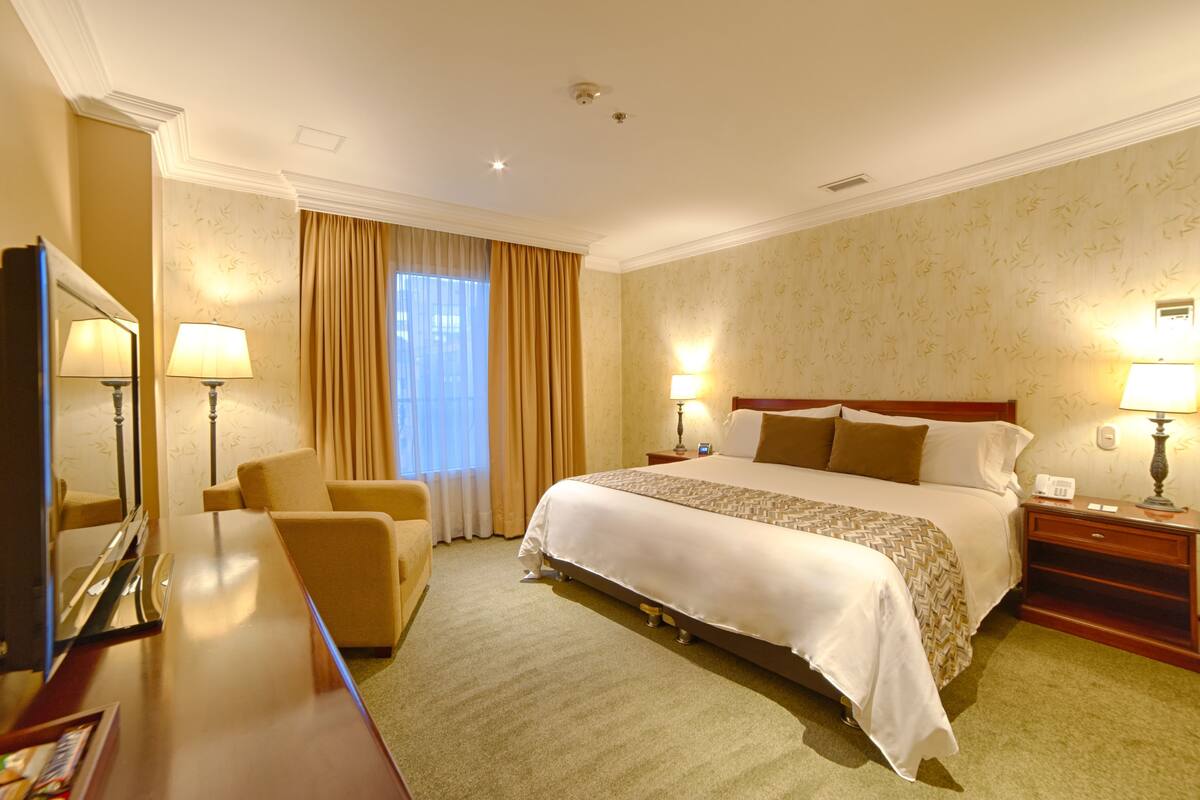 Una habitación de hotel con una cama grande, dos lámparas de noche, un escritorio con una silla, un espejo y una ventana con cortinas.