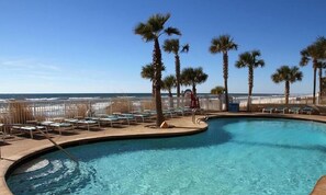 A swimming pool with lounge chairs and palm trees, surrounded by a fence and overlooking the ocean.