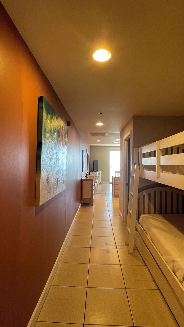 A hotel corridor with bunk beds on the right, a painting on the left, and a desk with a chair in the middle.