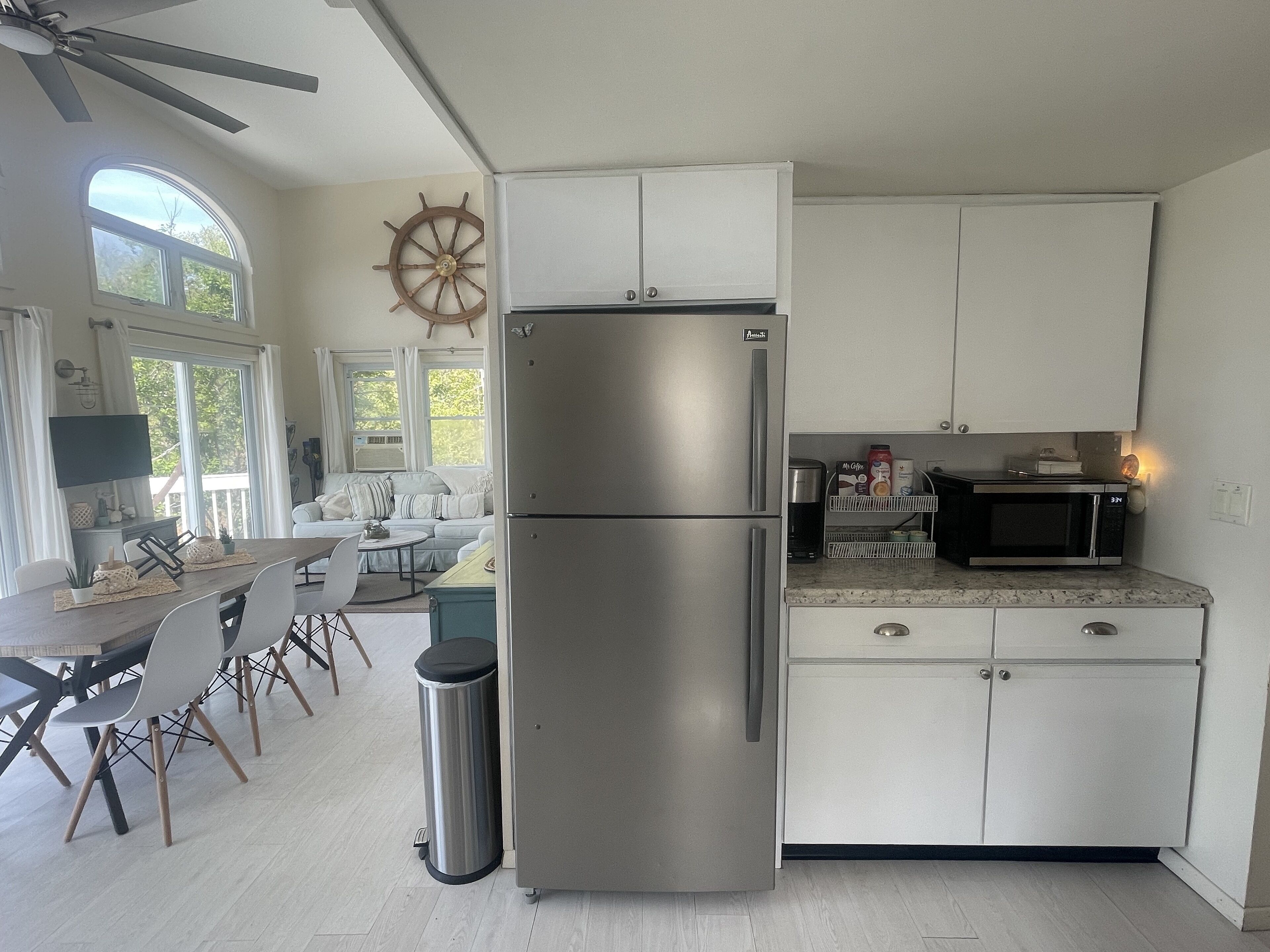 A modern kitchen with white cabinets, a stainless steel refrigerator, and a microwave. There is a dining area with a table and chairs, and a large window providing natural light.