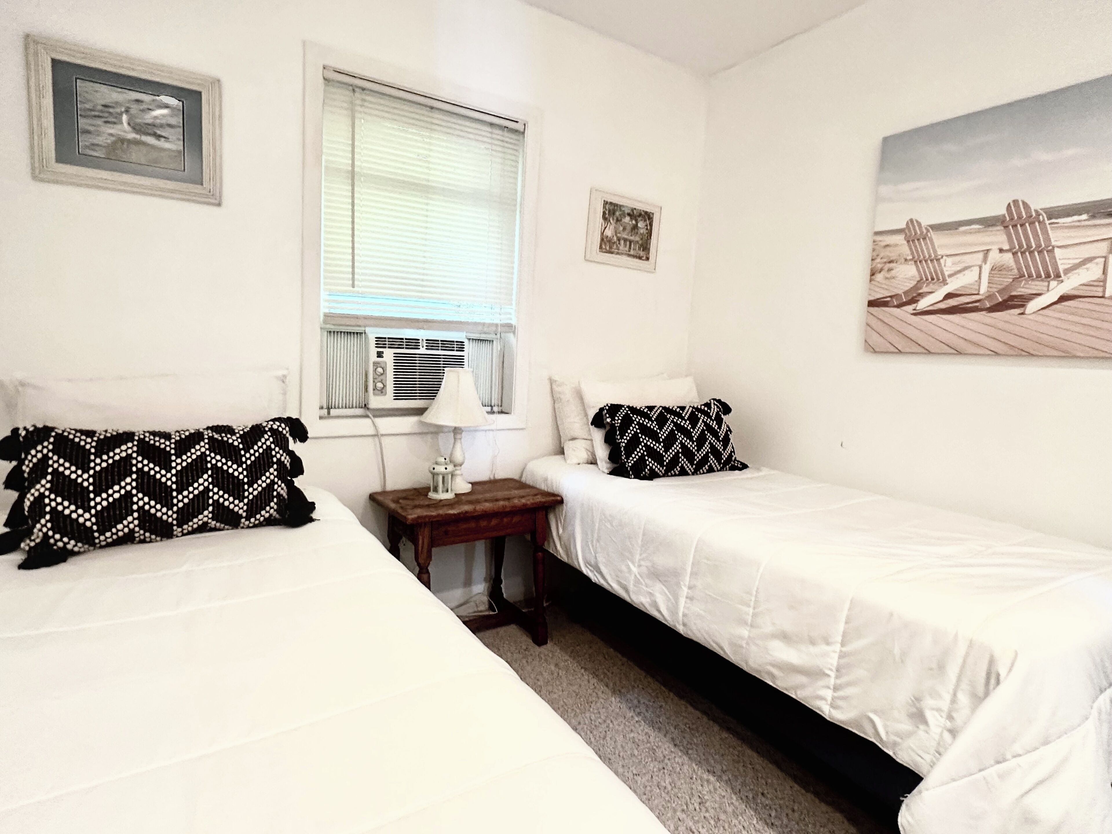 Une chambre avec deux lits, une fenêtre avec des stores, une petite table avec une lampe et un tableau sur le thème de la plage accroché au mur.