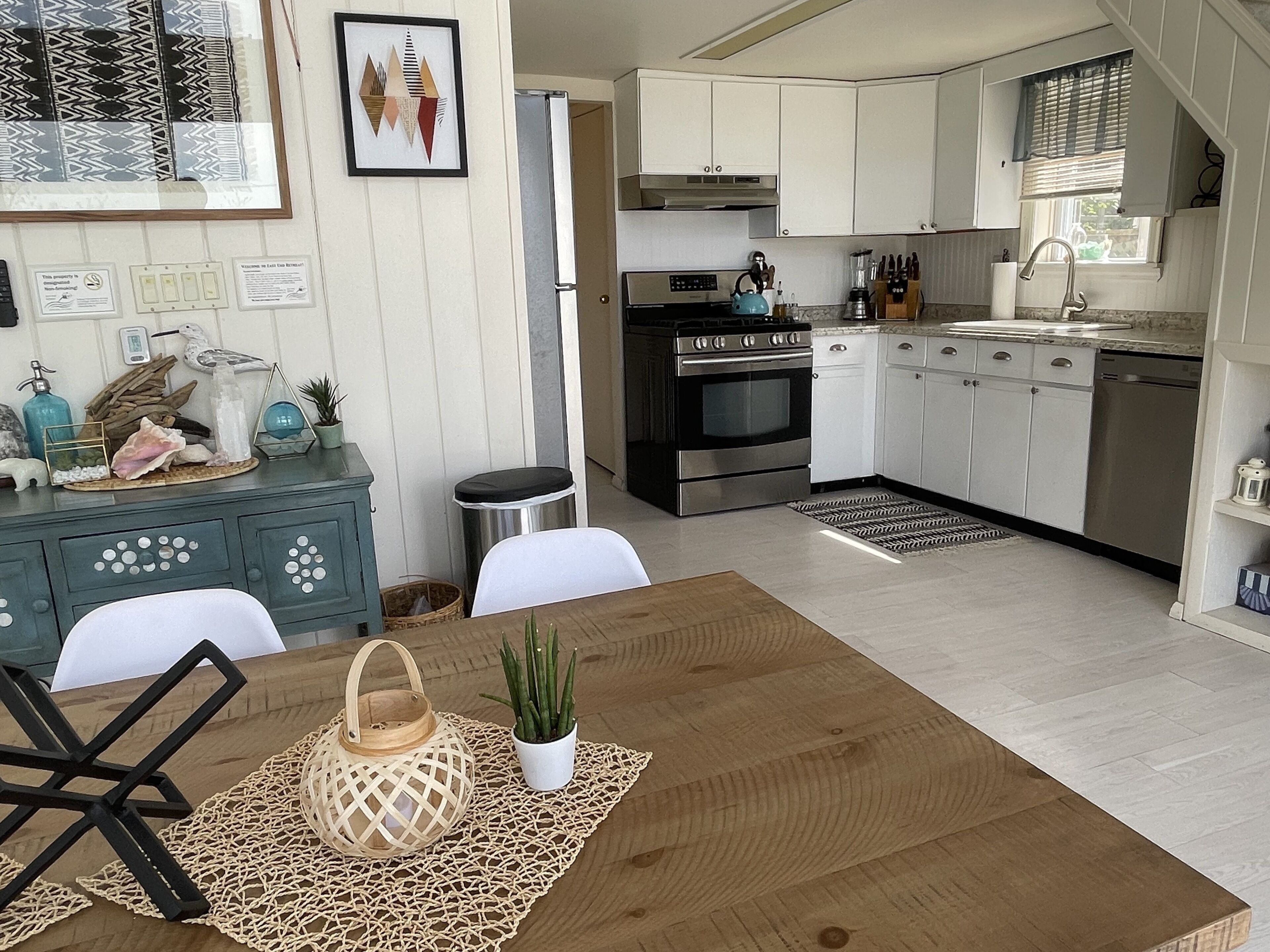 A modern kitchen with white cabinets, a stainless steel oven, and a dishwasher. There is a dining table with a placemat and a potted plant.