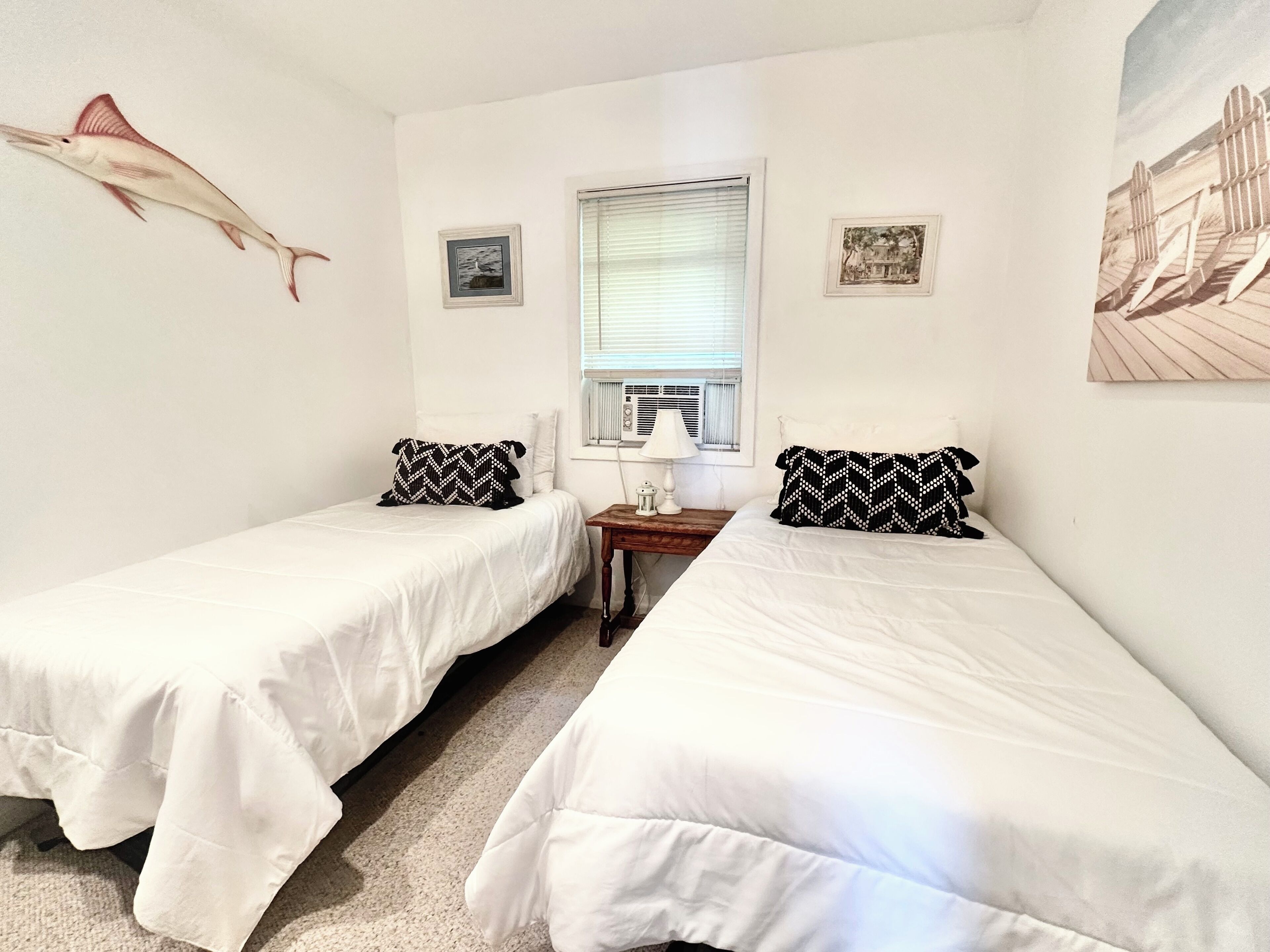 Quarto com duas camas de solteiro, um enfeite de peixe na parede, janela com persianas e duas imagens emolduradas.