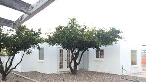A white building with two windows and a door, surrounded by gravel and trees. - Clean, renovated 1 Bedroom with everything you need. Granite, Travertine! (Tucson)