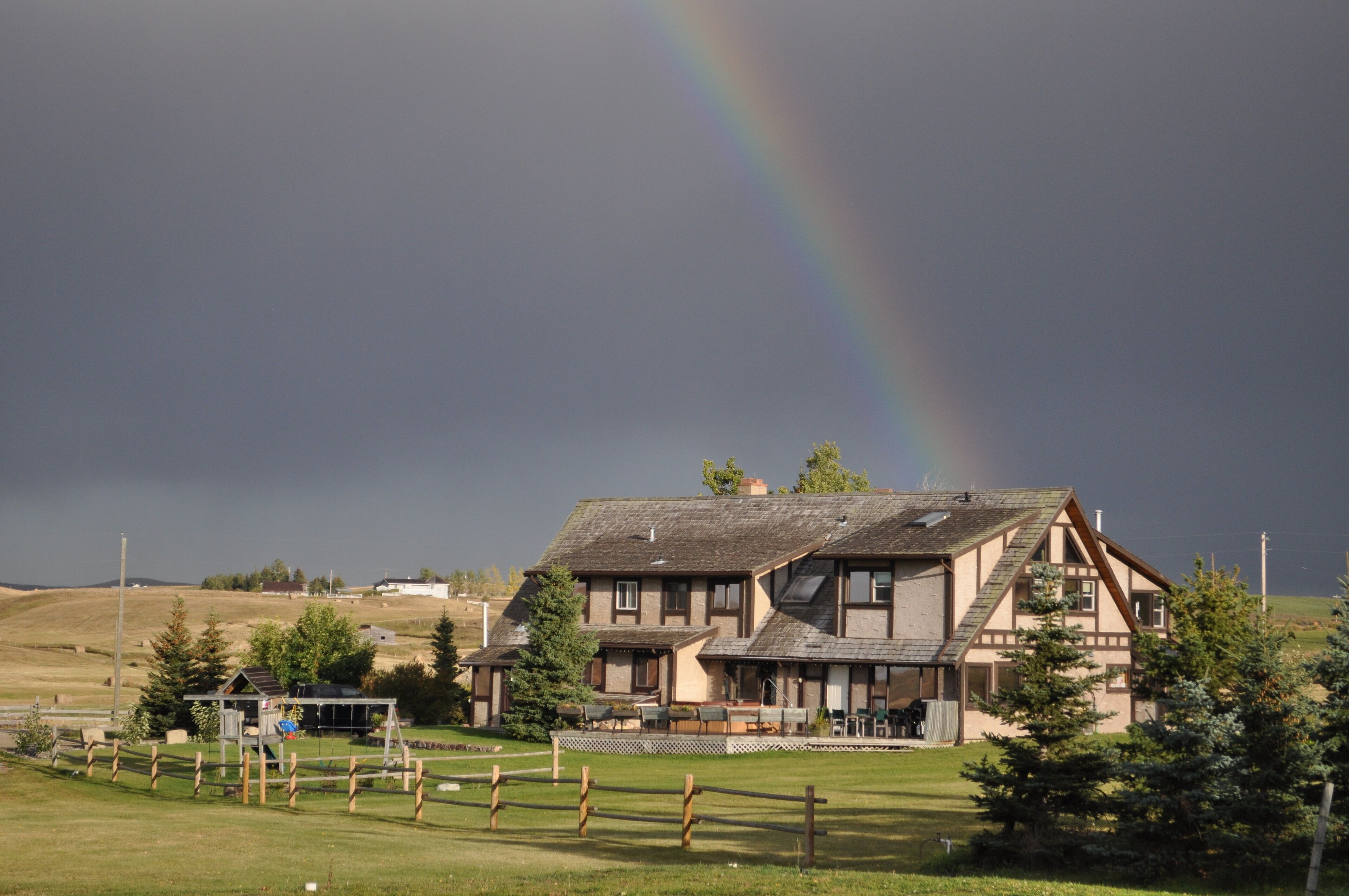 A two-story house with a sloped roof, a fenced yard, and a visible rainbow in the background.