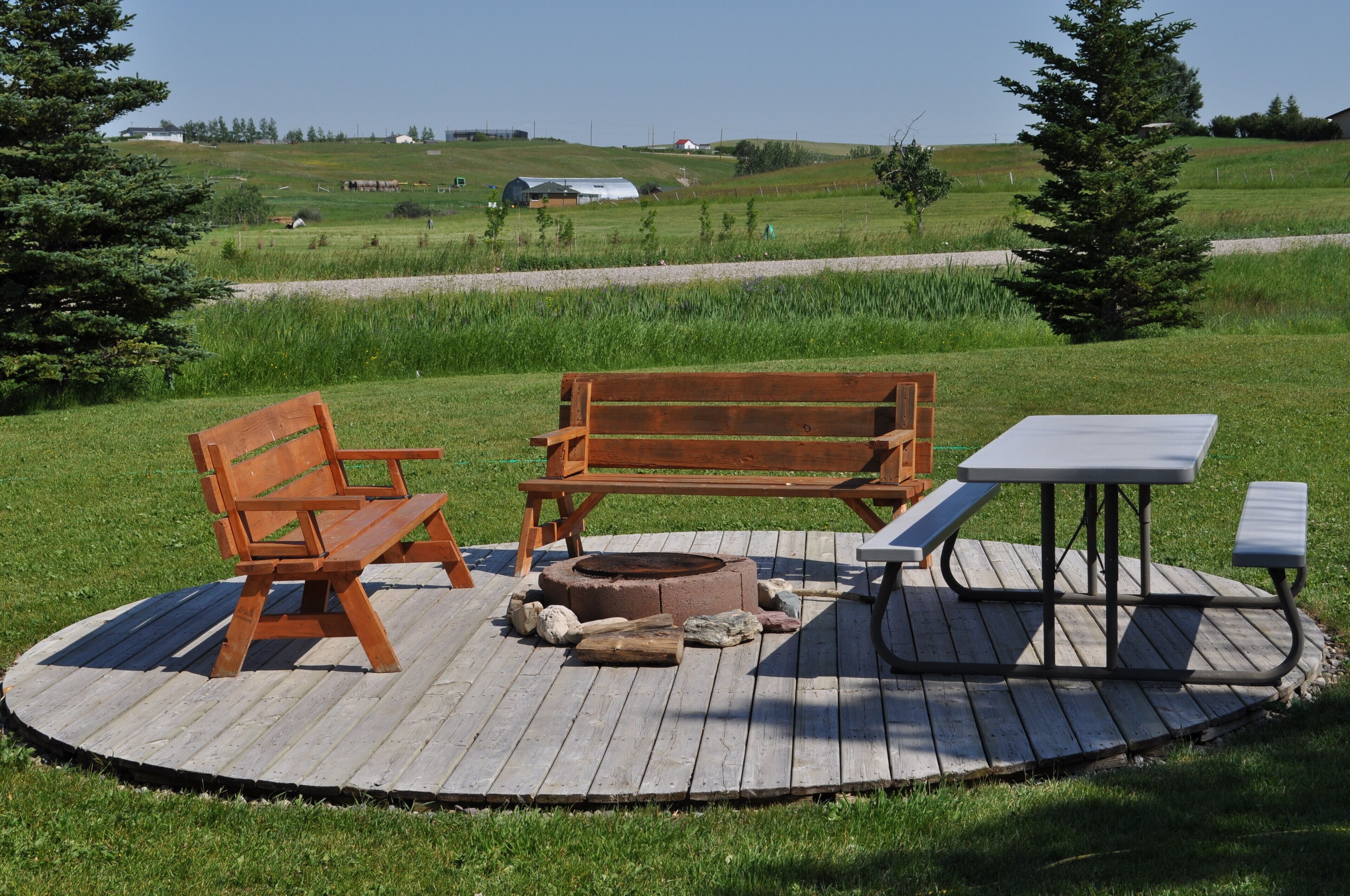 A wooden deck with a bench and picnic table, surrounded by a grassy field and trees.