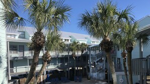 Area piscina con lettini e ombrelloni, circondata da palme e un edificio a più piani con balconi.