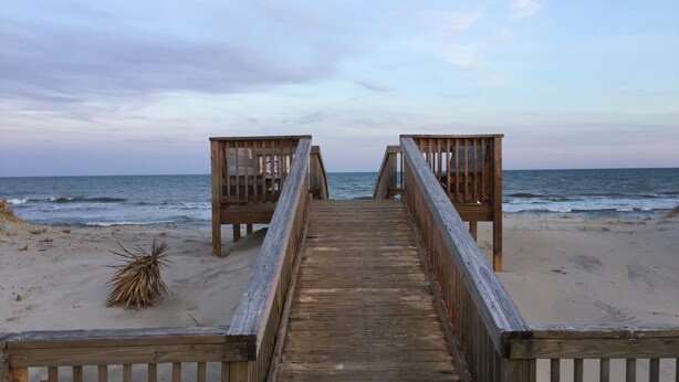 Eine Holzpromenade führt zu Strandliegen an einem Sandstrand, im Hintergrund das Meer.