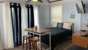 Ein Schlafzimmer mit einem Bett, einem Schreibtisch mit zwei Hocker, einem Deckenventilator, einem Fernseher und einem Fenster mit Jalousien.