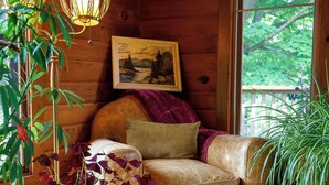 A cozy cabin interior with a leather sofa, a painting of a landscape, and a large window overlooking greenery. - Runaway Rooster B&B - The Coop (Prince Edward)