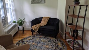 A living room with a dark sofa, a patterned rug, a small table with a plant, a bookshelf, and a framed picture on the wall. - Apt. retreat near lake and 215' waterfall, peaceful, lovely gardens.  (Trumansburg)