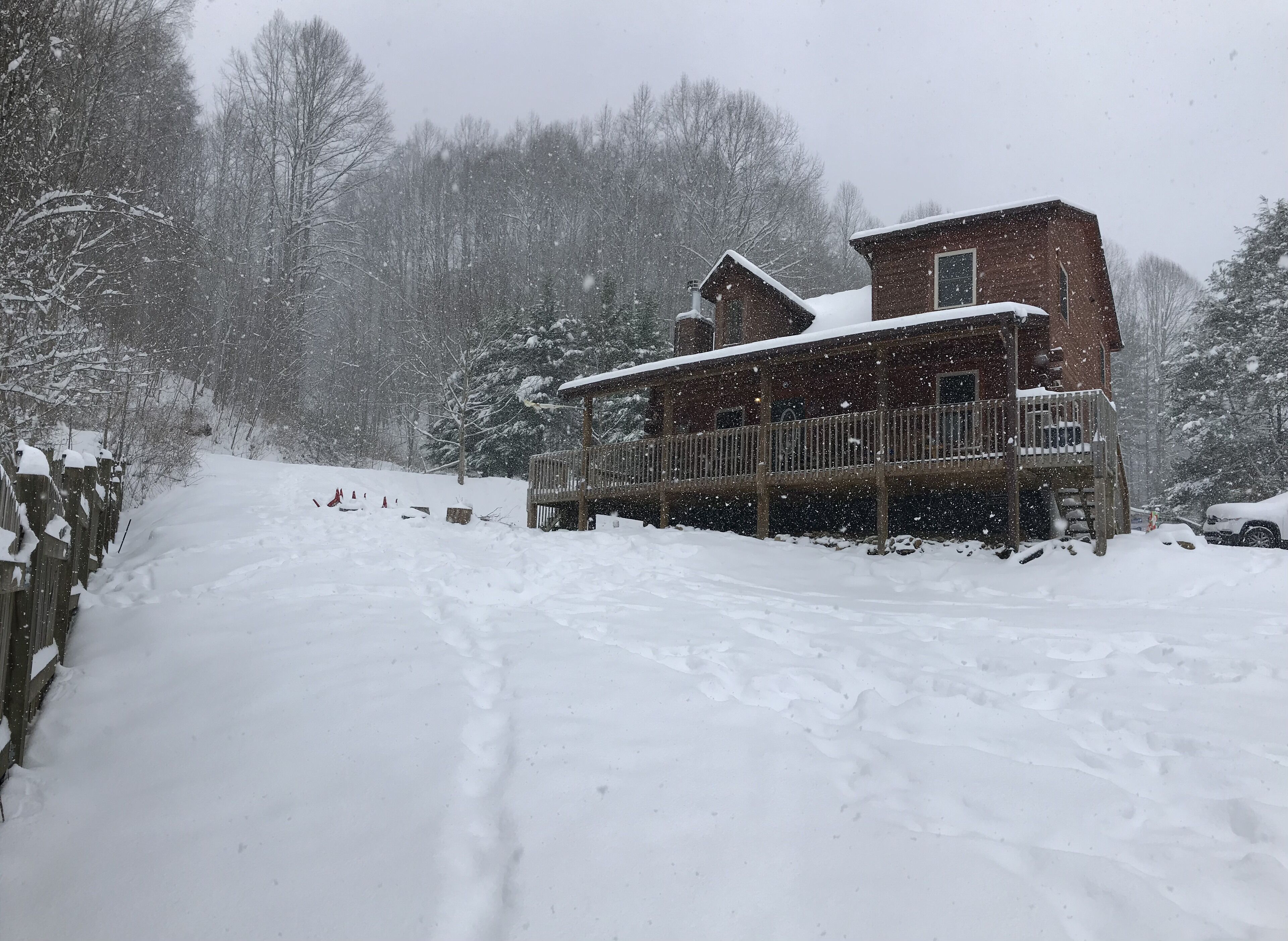 A wooden cabin with a balcony, surrounded by snow-covered trees and a snow-covered driveway.