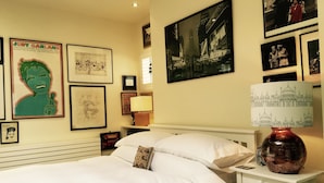 A bedroom with a bed, pillows, a nightstand, a lamp, and various framed pictures on the wall.