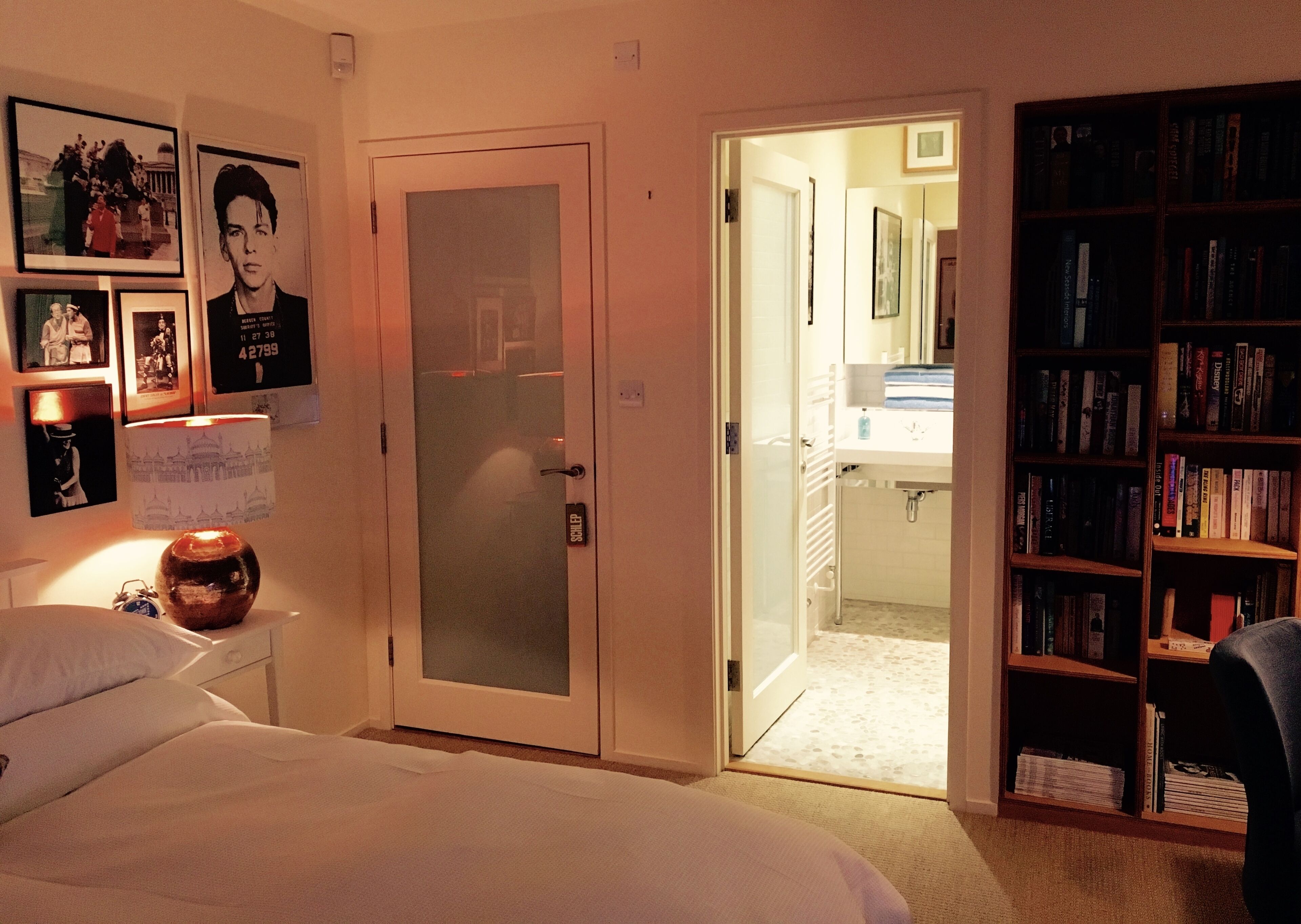 A bedroom with a bed, a bedside table with a lamp, a door leading to a bathroom, and a bookshelf filled with books.
