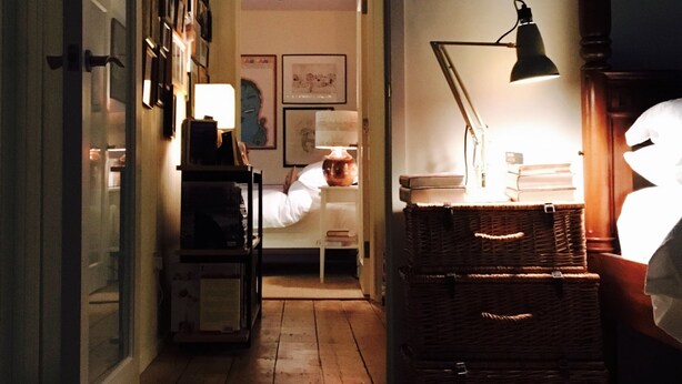A dimly lit room with a bed, a desk with a lamp, and a wicker chest of drawers.