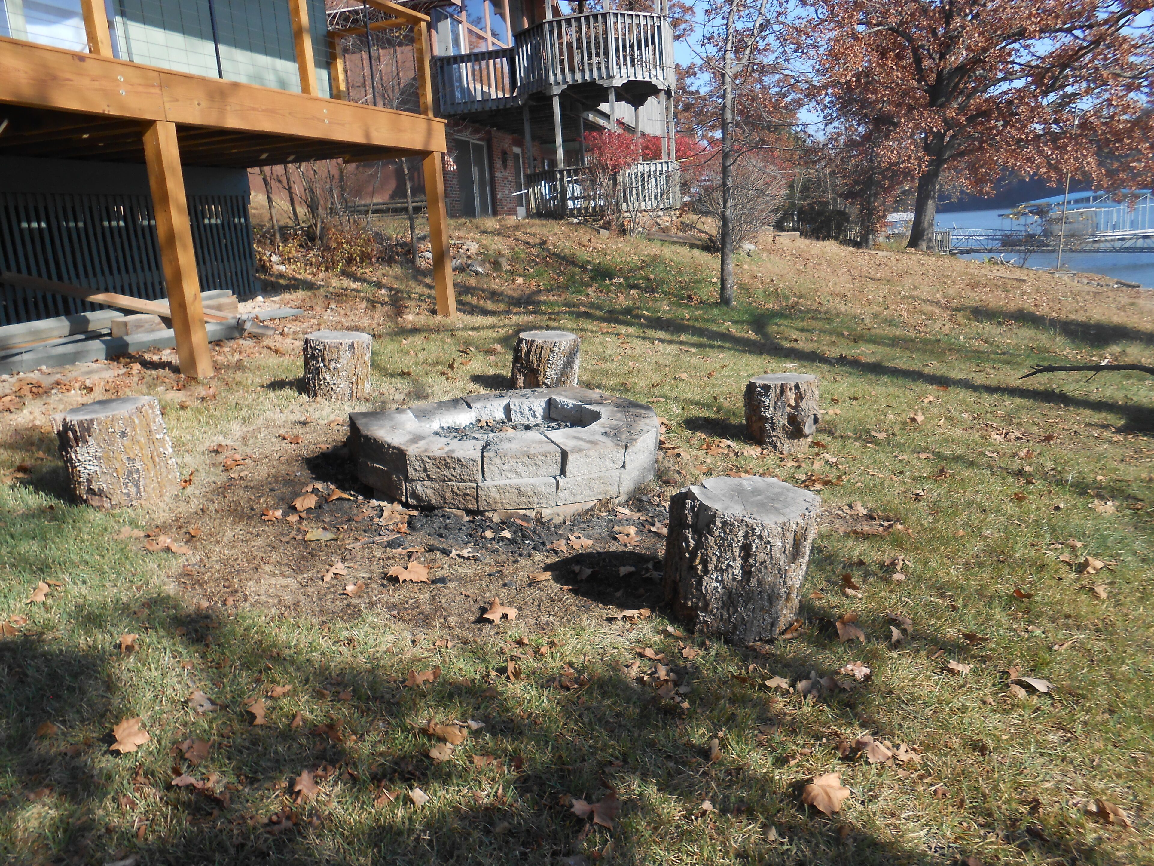A stone fire pit surrounded by tree stumps in a grassy area with a building in the background.