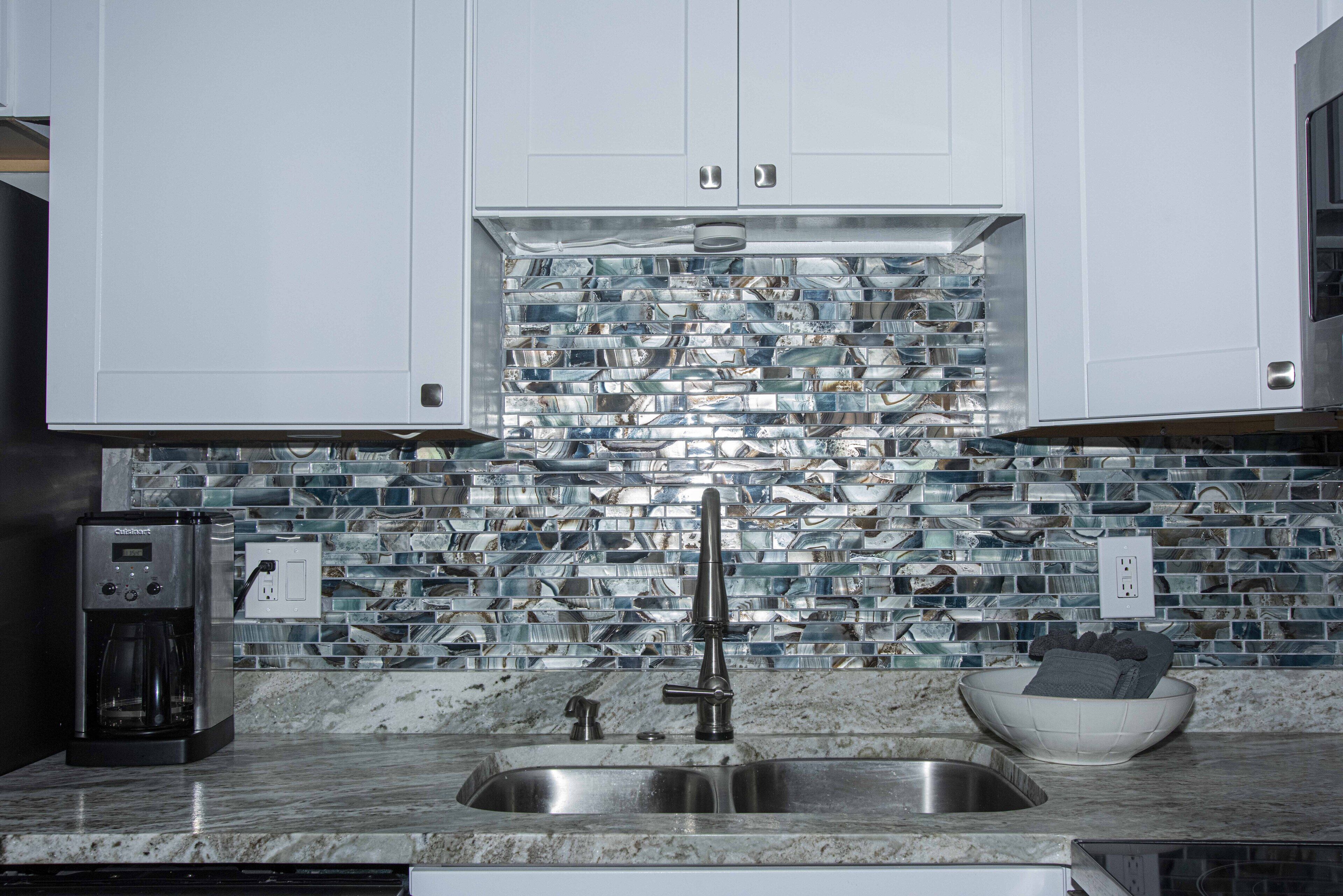 A kitchen with white cabinets, a marble countertop, a coffee maker, a sink, and a mosaic backsplash.