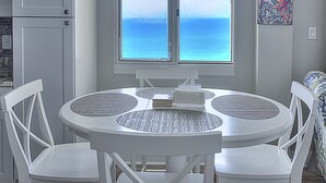 Una mesa de comedor redonda con cuatro sillas blancas, una ventana con vista al océano y un cartel de bienvenida colorido en la pared.