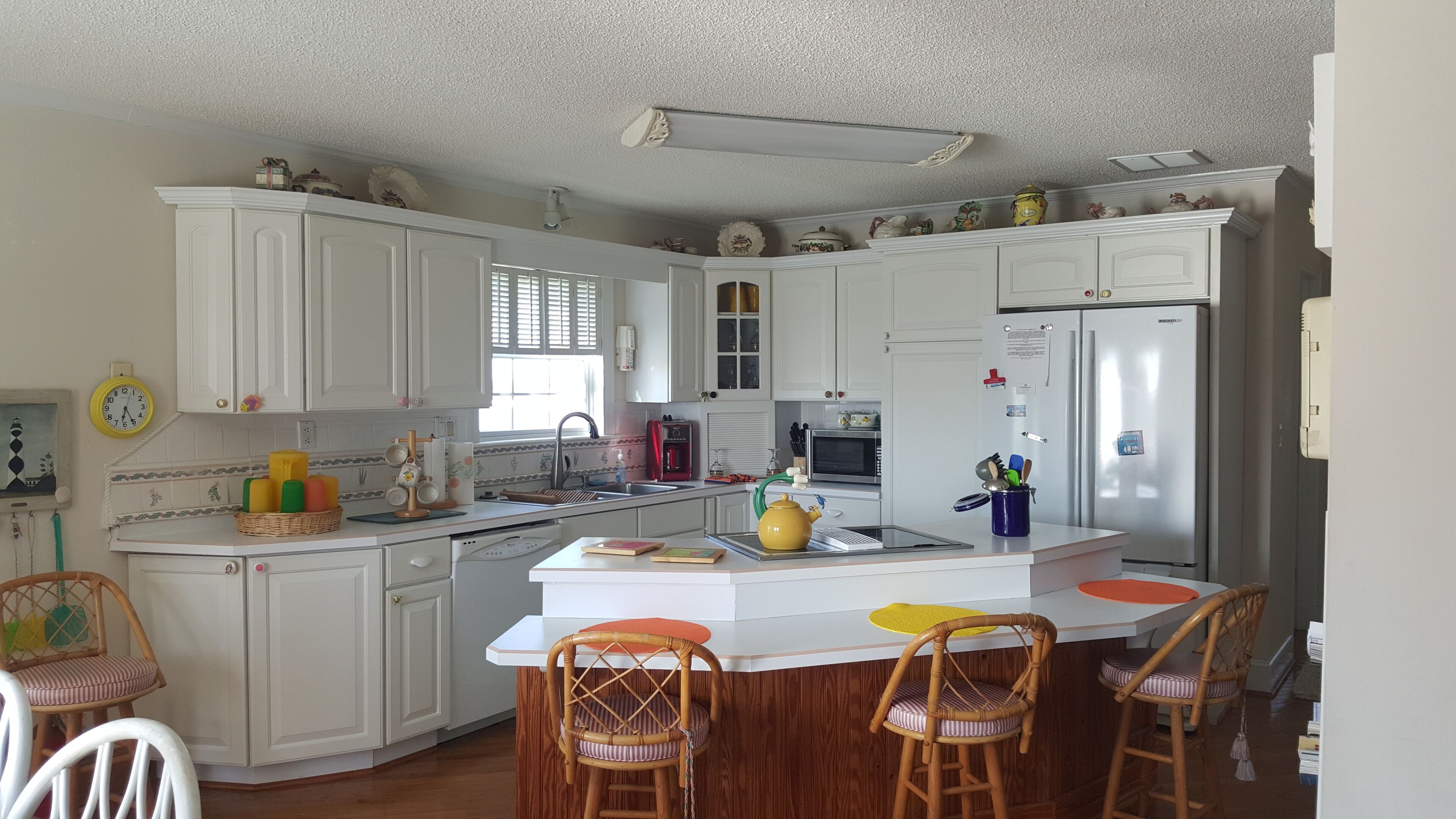 A kitchen with white cabinets, a refrigerator, a microwave, and a coffee maker. There are wicker chairs and a small island with a yellow kettle.