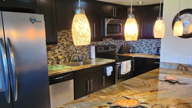 A modern kitchen with dark cabinets, a stainless steel refrigerator, a large island with a marble countertop, hanging pendant lights, and a clock on the wall. - Luxury Condominium In Key Largo... Sunsets, Marina (Key Largo)