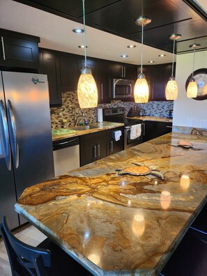 A modern kitchen with dark cabinets, a stainless steel refrigerator, a large island with a marble countertop, hanging pendant lights, and a clock on the wall. - Luxury Condominium In Key Largo... Sunsets, Marina (Key Largo)